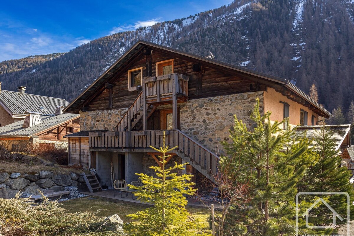 A charming stone and wood chalet with wooden stairs and balconies, nestled among pine trees against a snowy mountain backdrop under a clear blue sky.
