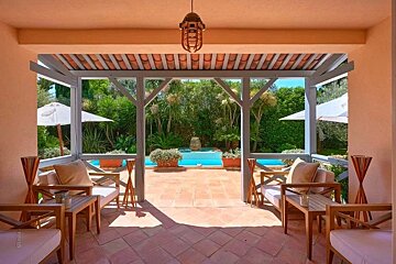 A Mediterranean-style covered patio with wooden furniture overlooking a swimming pool and lush garden.