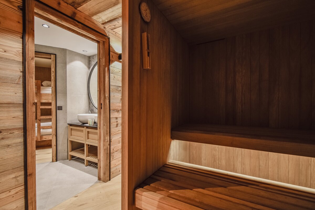 A cozy wooden sauna with tiered benches looking out into a modern bathroom vanity and bunk bed room beyond.