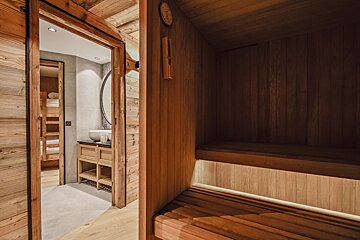 A cozy wooden sauna with tiered benches looking out into a modern bathroom vanity and bunk bed room beyond.