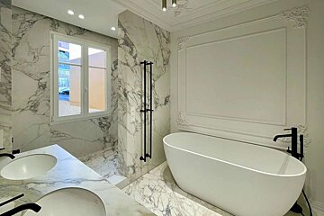Elegant bathroom with white marble throughout, double vanity, freestanding tub, and black fixtures. Features classic wall molding and a bright window.