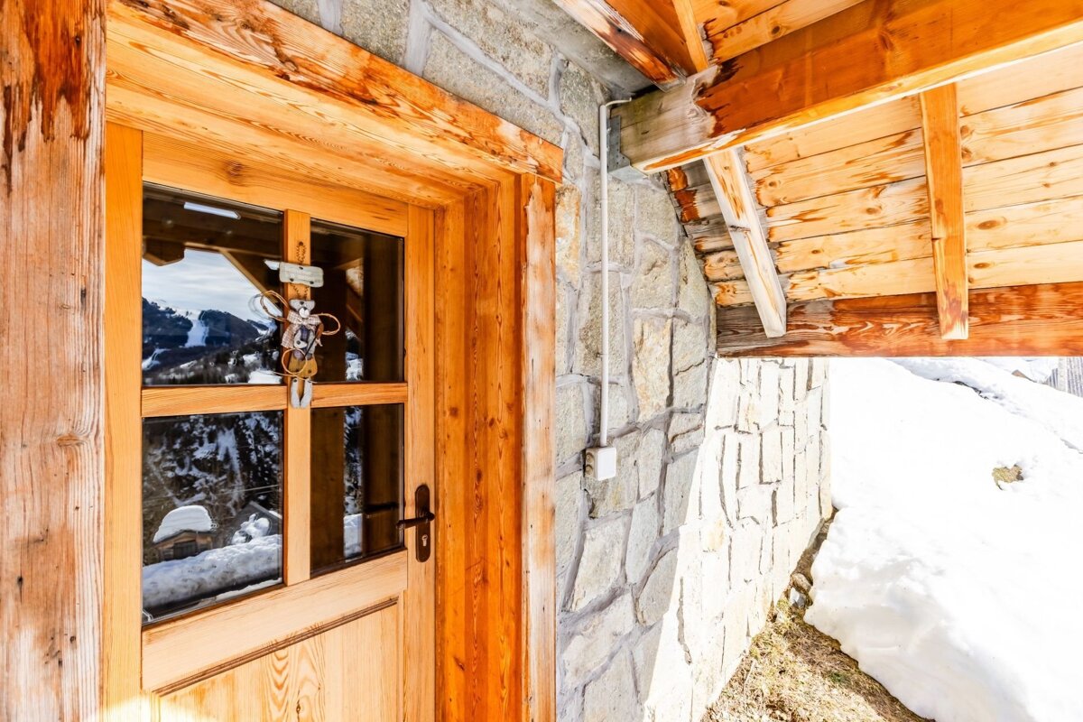 Rustic wooden door of a stone chalet in a snowy alpine setting, with mountains reflected in the glass.