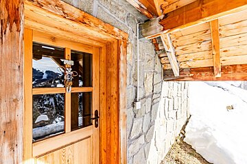 Rustic wooden door of a stone chalet in a snowy alpine setting, with mountains reflected in the glass.