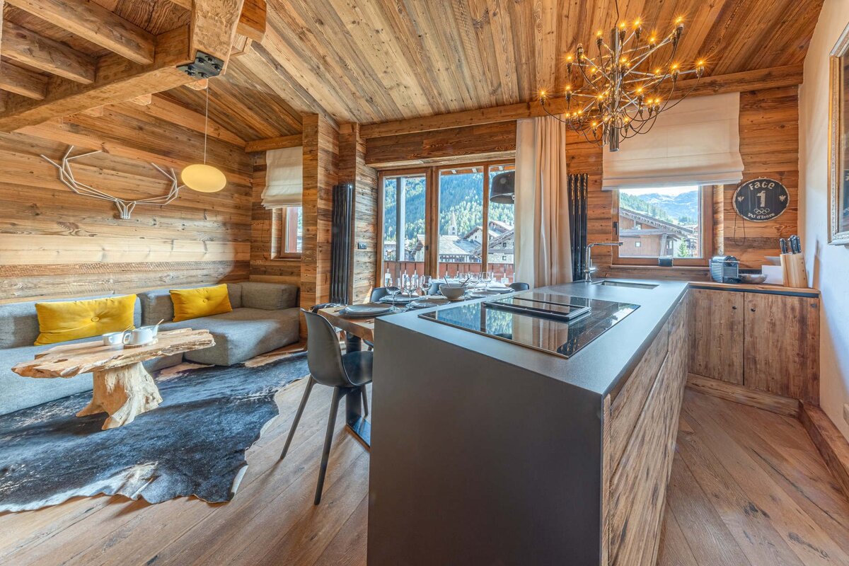A cozy, modern chalet interior featuring rich wooden walls, ceiling, and floors, an open kitchen/living area, and a scenic mountain view.