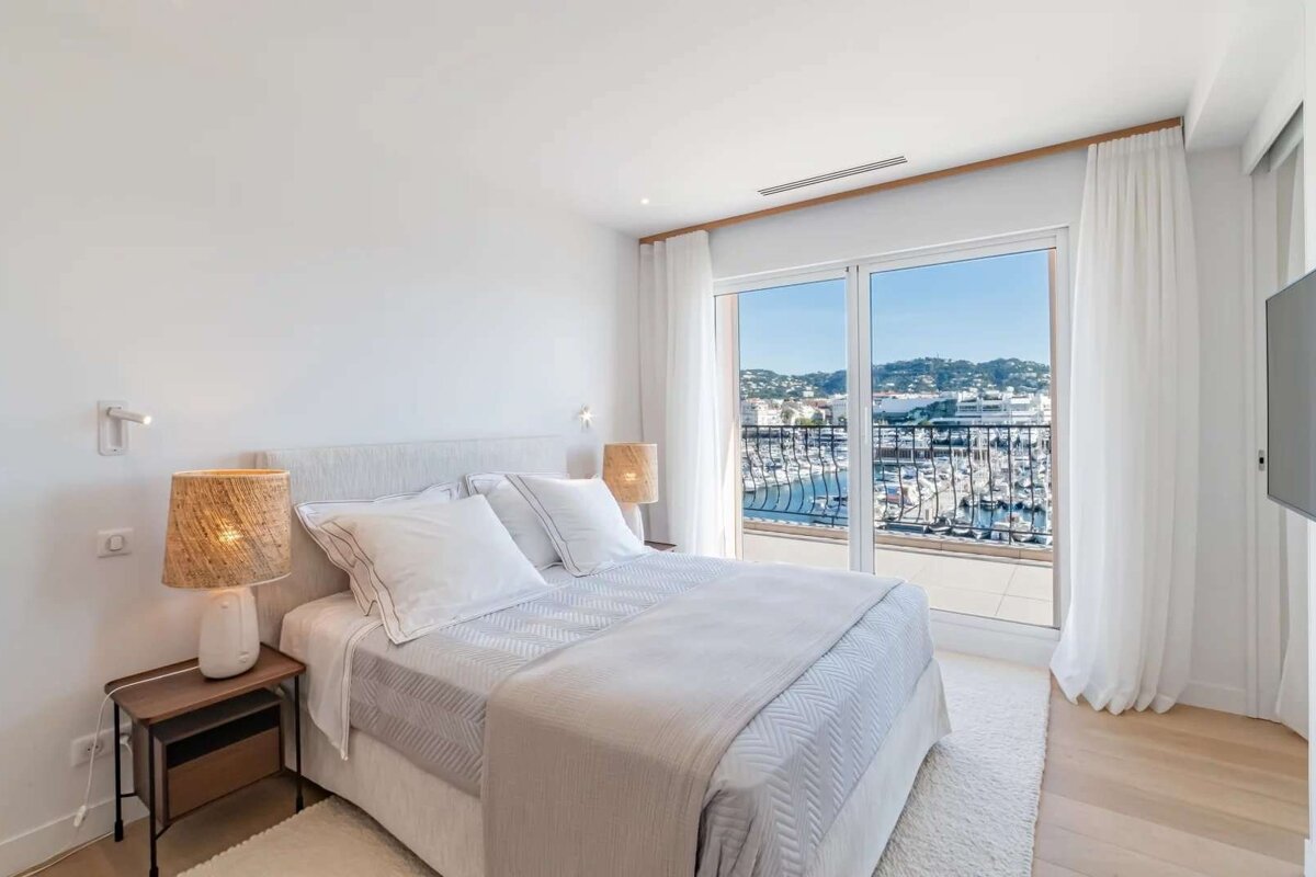 Bright, minimalist white bedroom with a balcony and large windows offering a stunning view of a sunny marina filled with boats and a coastal town.