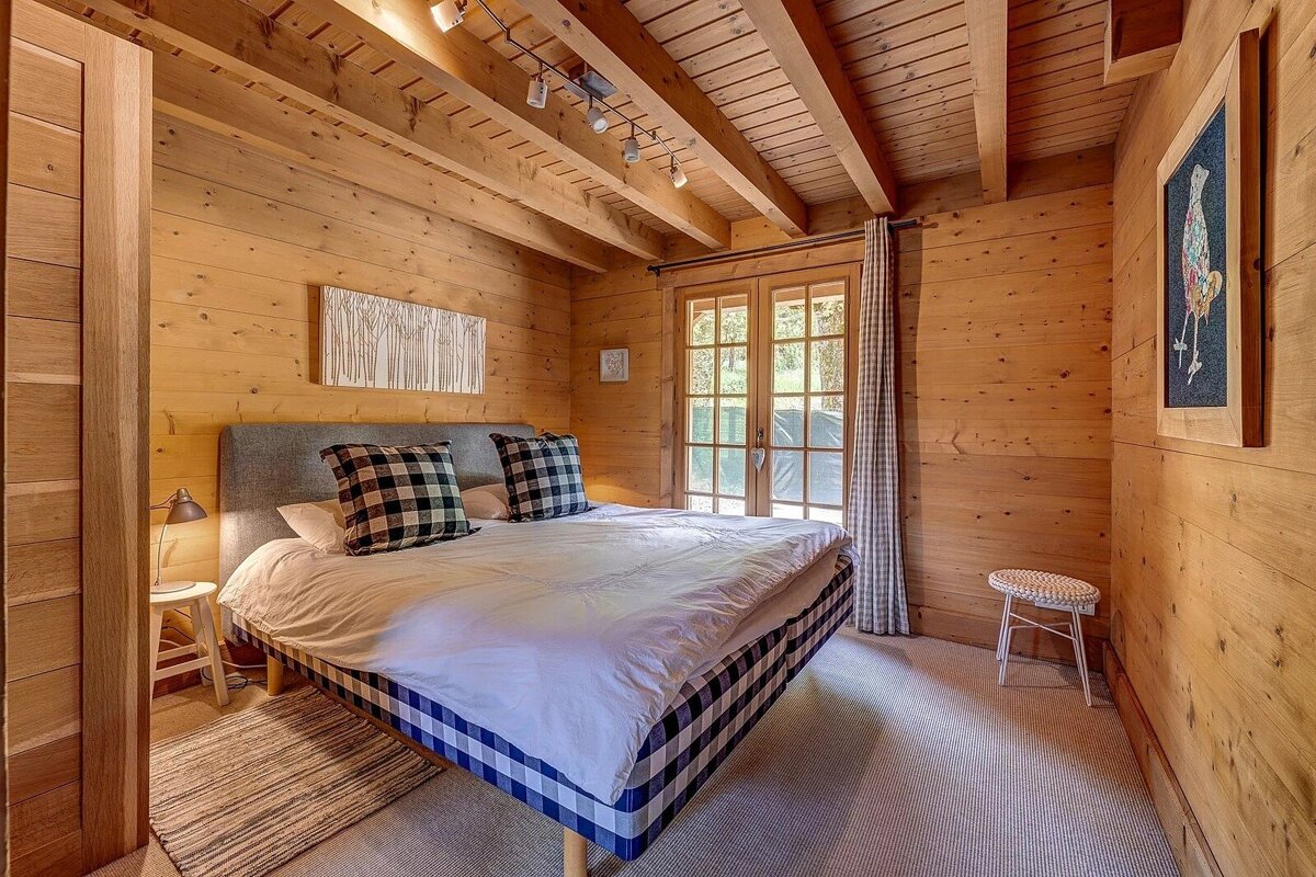 A cozy wooden cabin bedroom featuring a bed with blue plaid details, exposed ceiling beams, and glass doors letting in light.