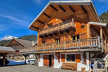 A traditional wooden alpine chalet with balconies, set against a backdrop of snowy mountains and a bright blue sky.