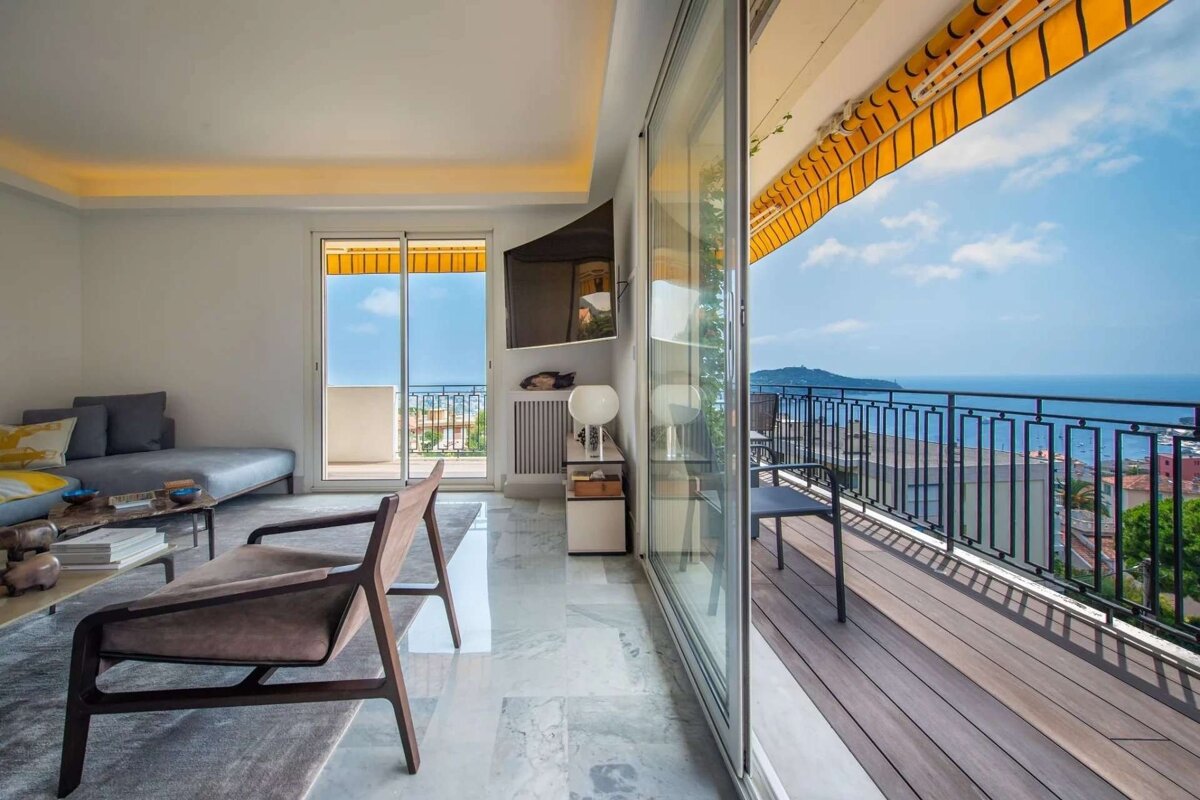 Modern living room with marble floors and a large balcony offering panoramic sea views under a clear sky.