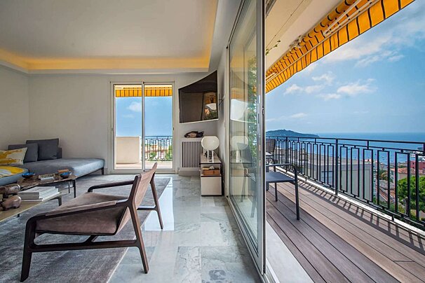 A bright, modern living room opens to a wide balcony with a stunning panoramic sea view, featuring large sliding glass doors and a vibrant yellow awning.
