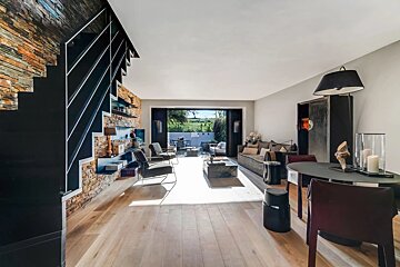 Sleek, modern open-plan living room with light wood floors, a stone wall, black staircase, and direct access to a furnished outdoor patio.