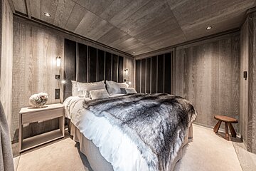 A modern, cozy bedroom with light wood paneling walls & ceiling. Features a large bed with a dark headboard and a plush faux fur throw.