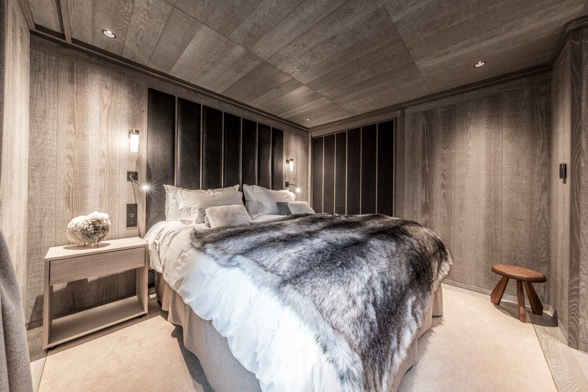 A luxurious, cozy bedroom with light wood paneling, a large bed featuring white linens and a plush faux fur throw, and a dark upholstered headboard.