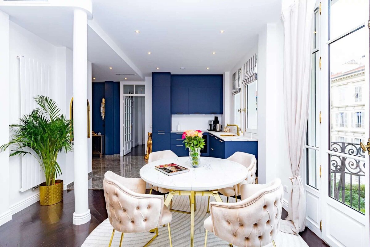 An elegant open-concept living space features a blue kitchen, white marble dining table with velvet chairs, and large windows leading to a balcony.