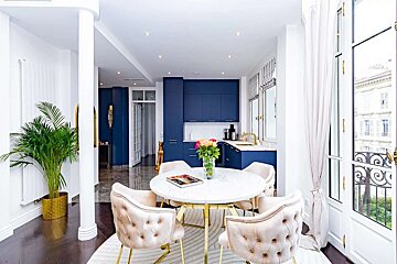 A chic, bright open-plan apartment featuring a navy kitchen, white marble dining table, velvet tufted chairs, and elegant gold accents.