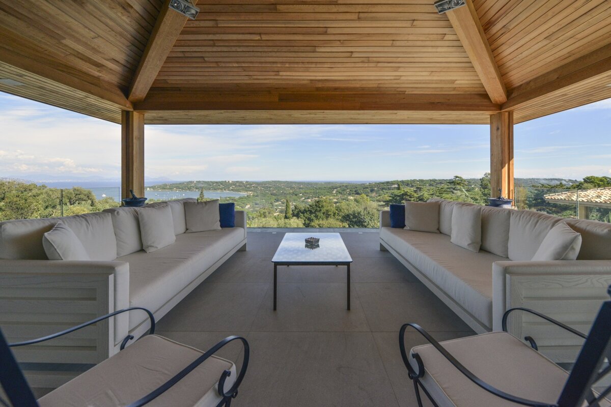 A luxurious open-air patio with a wooden ceiling, modern furniture, and a stunning panoramic view of a coastal bay, sea, and lush green landscape.