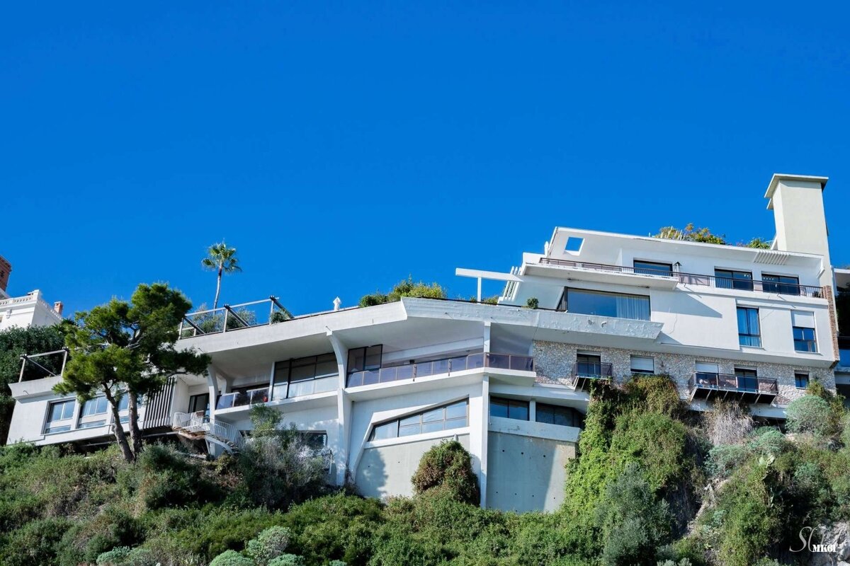 A modern, multi-level white house with balconies and large windows built into a lush green hillside under a clear blue sky, with a palm tree on top.