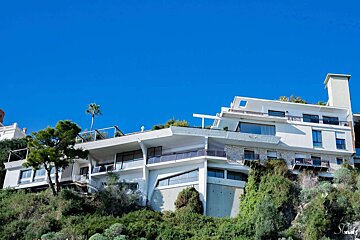 A modern, multi-level white house with balconies and large windows built into a lush green hillside under a clear blue sky, with a palm tree on top.