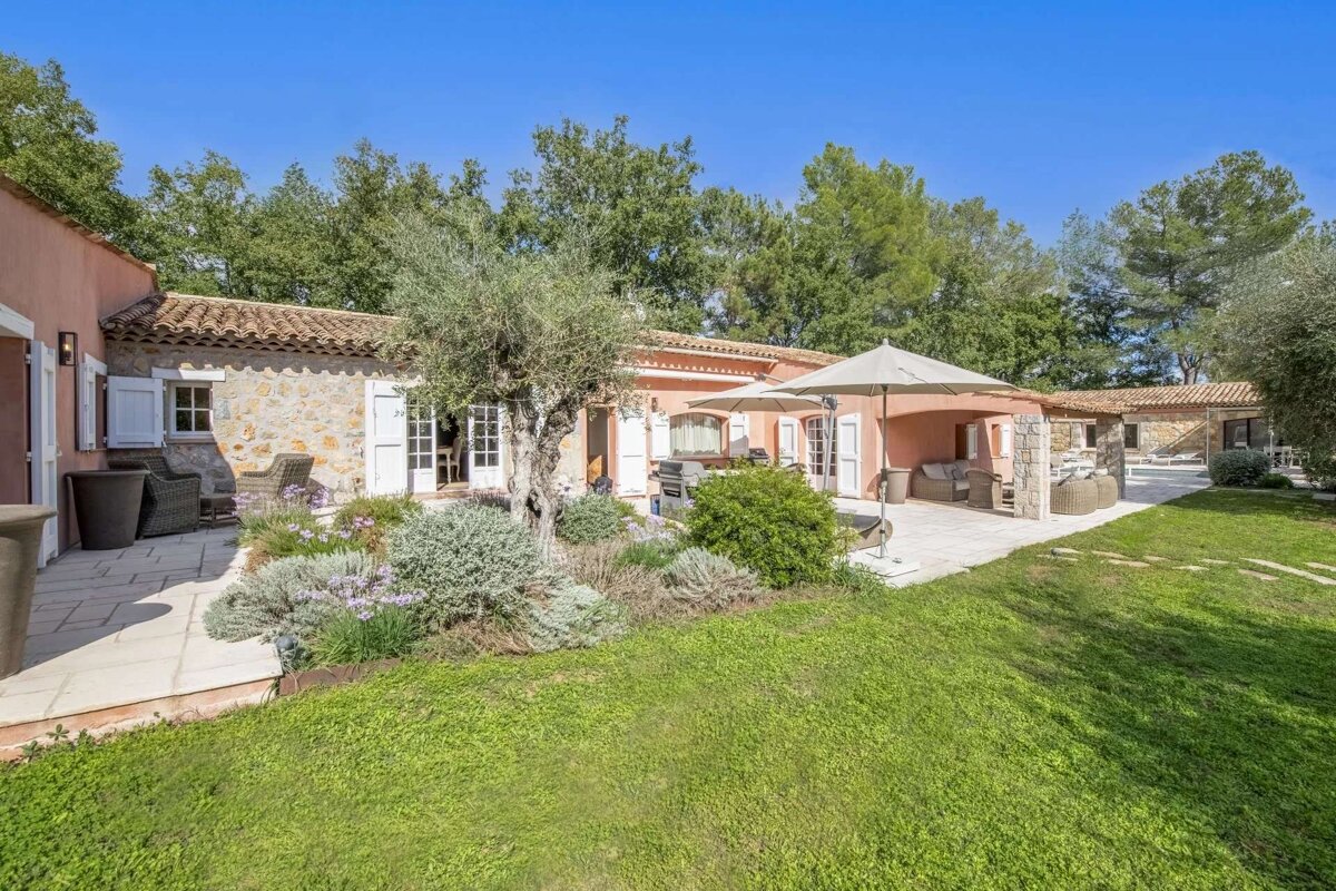 A charming peach and stone villa with lush gardens, shaded patios featuring outdoor furniture and umbrellas, and an olive tree under a bright blue sky.