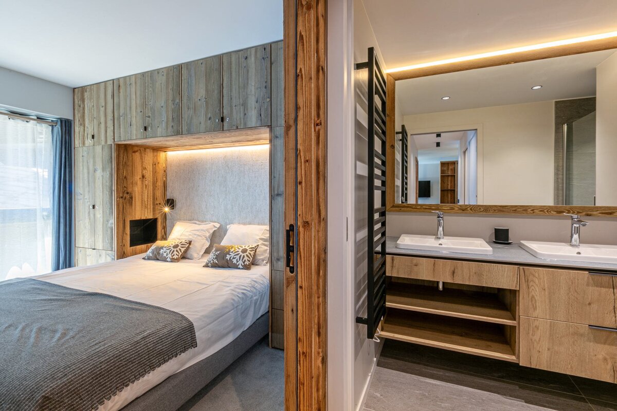 A cozy bedroom with a built-in wooden bed and storage, seamlessly connected to a modern bathroom with a large mirror and double vanity.