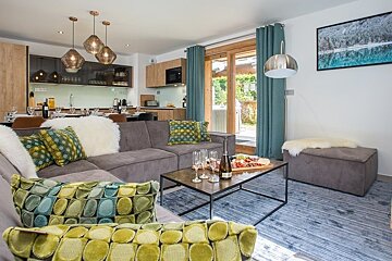 Cozy, modern living room featuring a grey sectional sofa with patterned pillows, a chic coffee table, and an open kitchen in the background.
