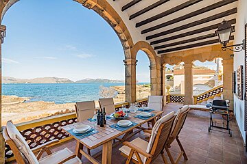 An arched seaside patio with a set dining table offers stunning views of the blue ocean and distant mountains under a clear sky.