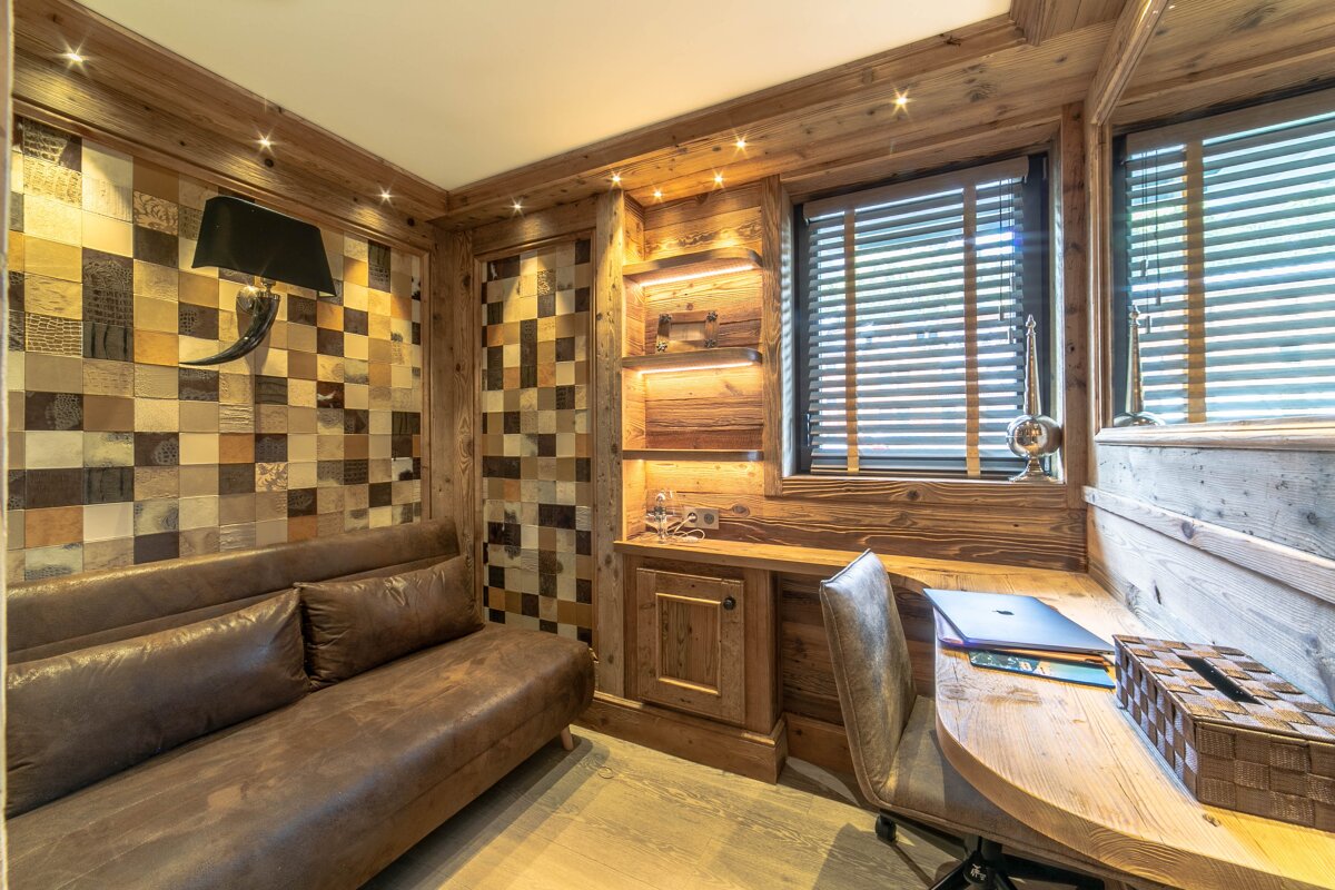 A cozy, rustic chalet-style room with extensive wood paneling, a checkered accent wall, leather sofa, and a desk with a laptop.