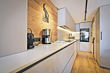 Modern minimalist kitchen with sleek white cabinets, a wood-paneled accent wall, coffee makers on the counter, and warm wooden floors.