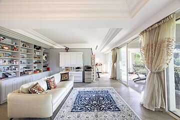 A bright, elegant living room with a white sofa, large bookshelves, a blue patterned rug, and sliding doors opening to an outdoor balcony.