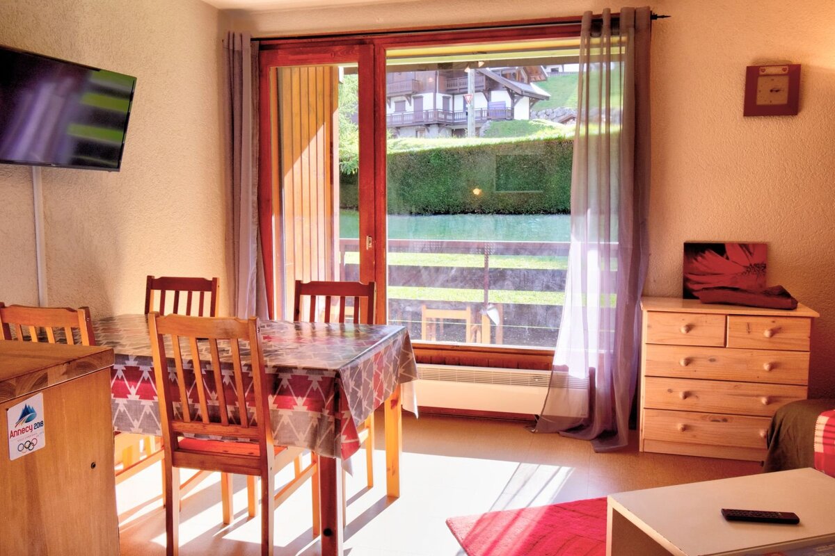 A bright room with a dining table, TV, and a large window offering a view of a charming village and lush green scenery under sunlight.