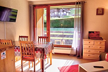 A bright room with a dining table, TV, and a large window offering a view of a charming village and lush green scenery under sunlight.