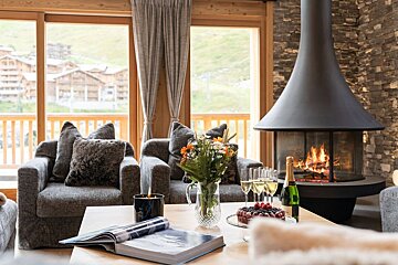 Cozy chalet living room with a modern fireplace, scenic mountain village view, and a coffee table with champagne, cake, and flowers.