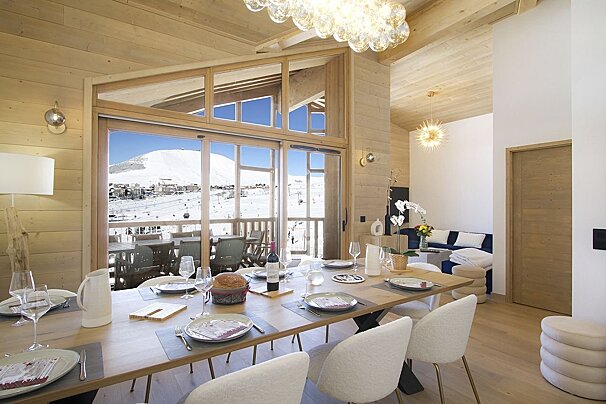 Chic dining room with a long table set for a meal, overlooking a stunning snowy mountain ski slope through expansive windows. Cozy, elegant chalet vibes.