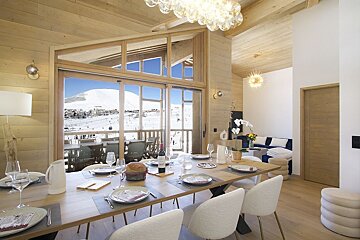 Chic dining room with a long table set for a meal, overlooking a stunning snowy mountain ski slope through expansive windows. Cozy, elegant chalet vibes.