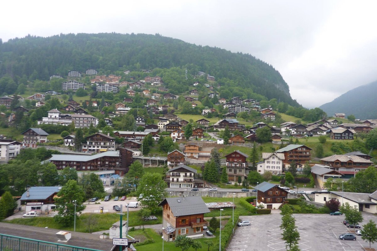 Alpine village with numerous chalet houses scattered across a lush green mountain slope under a cloudy sky.