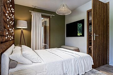 A modern bedroom with a white bed, olive green and wood panel walls, decorative string lights, a tripod lamp, wall-mounted TV, and wooden door.