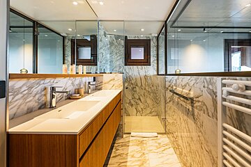 Luxurious modern bathroom with marble walls and floor, dual wood vanity, glass shower enclosure, and a window reflecting in mirrors.