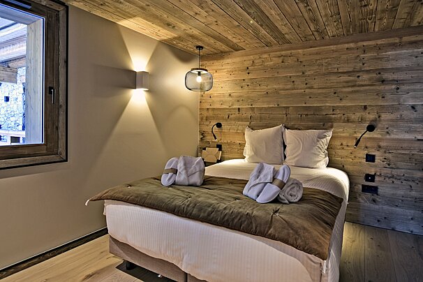 Cozy bedroom with wooden walls and ceiling. A bed with white linens, brown throw, towels, and slippers. Warm lights and a window.