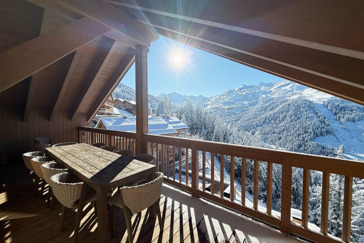 Wooden chalet balcony with a dining table overlooking a breathtaking, sunlit snowy mountain landscape.