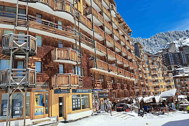 Sunny ski resort with multi-story wooden buildings and balconies, set against snow-covered mountains under a clear blue sky. Shops line the snowy street.