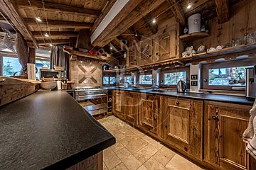 A rustic, cozy wooden kitchen with exposed beams, dark countertops, stainless steel appliances, and multiple windows looking out to the outdoors.
