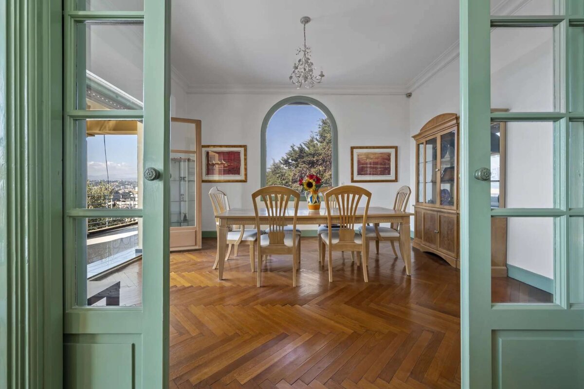 An elegant dining room with a wooden table, herringbone floor, and an arched window overlooking trees. Green-framed doors open to a balcony with city views.