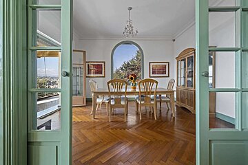 An elegant dining room with a wooden table, herringbone floor, and an arched window overlooking trees. Green-framed doors open to a balcony with city views.
