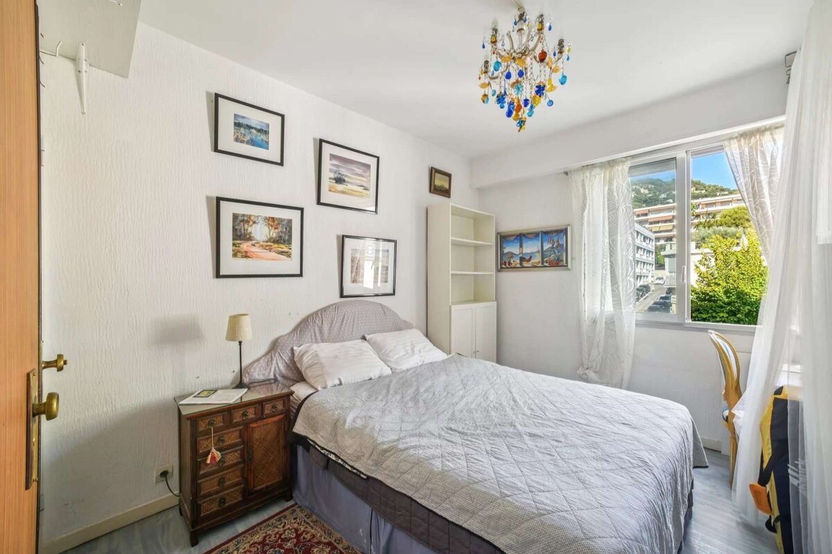 A bright bedroom with a bed, white walls, multiple framed pictures, a colorful chandelier, and a window overlooking a cityscape with green hills.