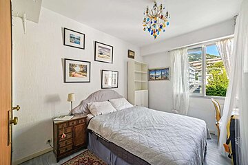 A bright bedroom with a bed, white walls, multiple framed pictures, a colorful chandelier, and a window overlooking a cityscape with green hills.