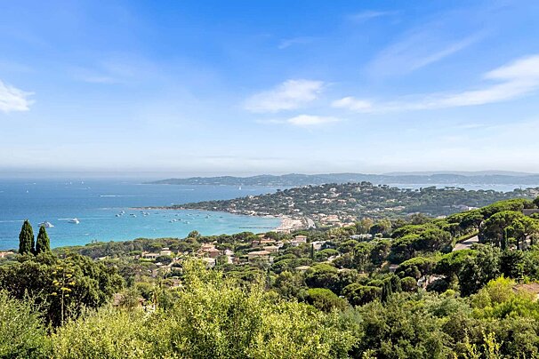 Aerial view of a vibrant coastal town and bay with boats, sandy beaches, and lush green hills under a clear blue sky, evoking a Mediterranean scene.