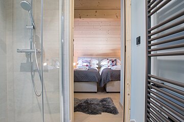 View from a modern bathroom shower into a cozy bedroom with two beds, light wood paneling, a fluffy grey rug, and a towel radiator.