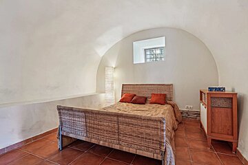 Minimalist bedroom with arched white ceiling, wicker bed frame, terracotta tiled floor, and a small, barred window above the bed.