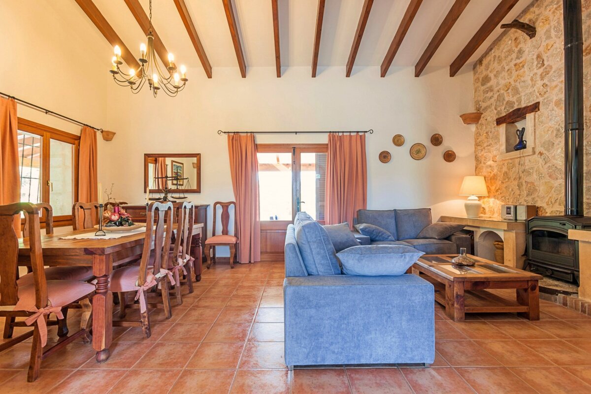 Cozy rustic living/dining room with exposed beams, terracotta floor, stone wall, fireplace, wooden table, and blue sofas.