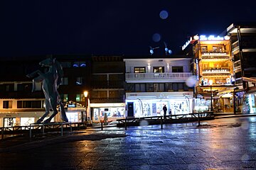 Night scene of a brightly lit street in a resort town with shops, a horse statue, and wet ground reflecting colorful lights.