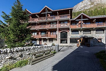 A multi-story alpine building with wood balconies and a stone base, an evergreen tree, and mountains in the background under a blue sky.
